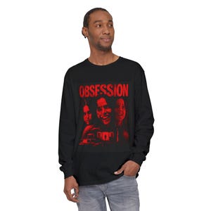 Puede incluir: Una camiseta negra de manga larga con un estampado gráfico rojo en el pecho. El diseño muestra la palabra OBSESSION en letras mayúsculas negritas sobre una ilustración estilizada de tres rostros y una casa, todo en un tono rojo monocromático.