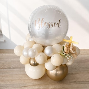 May include: A balloon arrangement featuring a large clear bubble balloon with the word Blessed printed in gold script. The base consists of various cream, white, and metallic gold balloons, accented with small white faux flowers and a gold cross.