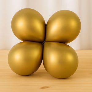 May include: Four gold-colored balloons arranged in a cloverleaf shape. The balloons have a metallic sheen and are resting on a light-colored wooden surface. The background is a soft, neutral color.