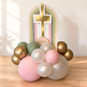 May include: A decorative centerpiece featuring a vertical arch-shaped sign with a gold cross and the name EMMA printed in gold. The sign is surrounded by a cluster of balloons in shades of sage green, metallic gold, soft pink, and white.