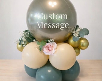 May include: A balloon arrangement featuring a large silver orb balloon with the text "Custom Message". The arrangement includes cream, gold, and teal balloons, accented with greenery and a pink rose. A decorative centerpiece for celebrations.
