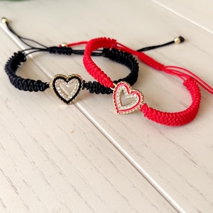 May include: Two friendship bracelets with heart-shaped charms. One bracelet is black with a silver and clear heart charm, and the other is red with a gold and clear heart charm. The bracelets are on a white wooden surface.