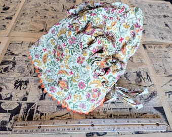 Puede incluir: Un gorro blanco de algodón con estampado floral en tonos rosa, amarillo y verde. El gorro tiene un borde festoneado con un ribete naranja.