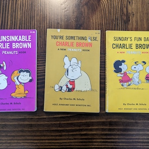 Könnte beinhalten: Drei Vintage-Taschenbücher mit den Figuren der Comicserie Peanuts. Die Bücher tragen die Titel "The Unsinkable Charlie Brown", "You're Something Else, Charlie Brown" und "Sunday's Fun Day, Charlie Brown". Jedes Buch zeigt eine andere Illustration von Charlie Brown und seinen Freunden.