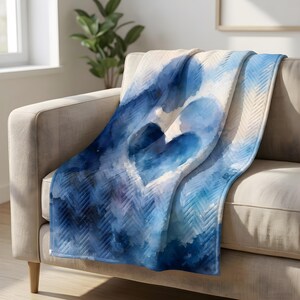 Può includere: Una coperta blu e bianca con un motivo a cuore ad acquerello drappeggiata su un divano beige. La coperta presenta un motivo a spina di pesce e un grande cuore astratto nei toni del blu.