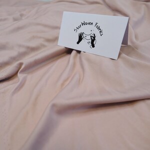 Könnte beinhalten: Nahaufnahme eines weichen, rosafarbenen Stoffes mit sichtbaren Falten und einer rohen Kante. Eine weiße Karte mit dem Text "Star Woven Fabrics" und einem Logo mit Händen, die Sterne halten, ist auf dem Stoff platziert.