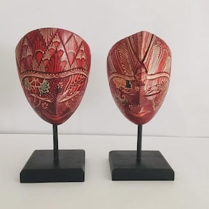 Op de afbeelding: Twee rode, gesneden houten maskers met ingewikkelde ontwerpen, tentoongesteld op zwarte standaards. De maskers hebben een traditionele esthetiek, met gedetailleerde patronen en een glanzende afwerking. De achtergrond is effen wit.