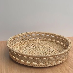 May include: A round, natural-toned woven rattan tray. The tray features an openwork design around the rim and a tightly woven base. The tray is sitting on a wooden surface.
