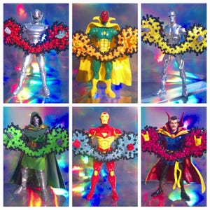 May include: Six action figures of Marvel Comics characters, including Ultron, Vision, and Iron Man, each adorned with a colourful, beaded garland. The figures are posed against a shimmering, iridescent background.