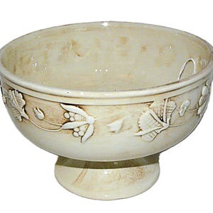 May include: Cream-colored ceramic bowl with a pedestal base. The bowl features embossed floral and leaf designs around the exterior. The interior of the bowl has a slightly darker, aged appearance.