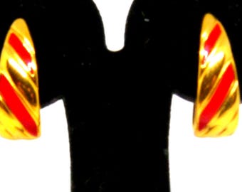 May include: A pair of gold-toned earrings with a curved design. Each earring features three diagonal red stripes. The earrings are displayed on a black stand against a white background.