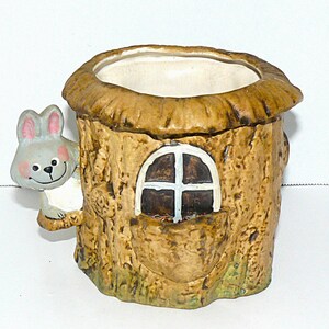 Peut inclure: Un pot de fleurs en céramique en forme de tronc d'arbre, avec une fenêtre ronde et un lapin gris souriant. Le pot a un extérieur marron clair avec un motif d'écorce texturée et un intérieur blanc.