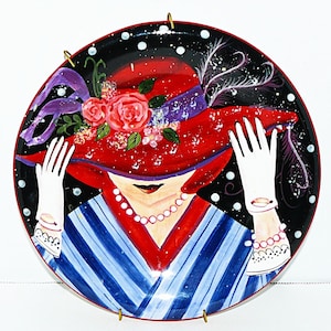 May include: Decorative plate depicting a woman in a red hat adorned with roses, purple ribbon, and white gloves. The plate has a black background with white polka dots and a red border. The woman's dress is blue and white striped.