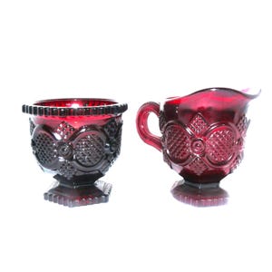 May include: A pair of ruby red glass vessels consisting of a sugar bowl and a creamer pitcher. Both items feature a pressed glass pattern with diamond-shaped textures, circular motifs, and a scalloped rim, all supported by a square pedestal base.