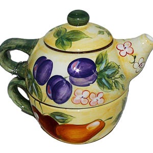 May include: A ceramic tea for one set featuring a yellow background with hand-painted purple plums, green leaves, small white flowers, and a large orange pear. The set includes a teapot with a green lid and a matching teacup stacked underneath.