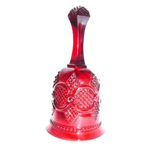 May include: A deep red glass bell featuring a textured diamond pattern across the body, circular embossed accents, and a smooth, tapered handle topped with a faceted, diamond-shaped finial. The base is finished with a beaded border detail.