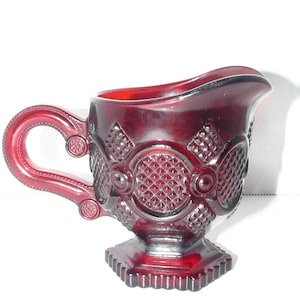 Pode incluir: Uma leiteira de vidro vermelho profundo com um padrão de diamante prensado dentro de medalhões circulares. O recipiente possui uma alça curva com detalhes decorativos, um bico largo e uma base quadrada com estrias verticais nas bordas.