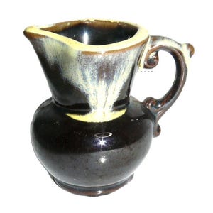 May include: A small ceramic creamer pitcher featuring a glossy dark brown glaze with a cream-colored drip pattern cascading from the rim. The pitcher has a rounded body, a wide pouring spout, and a curved handle with decorative scroll details.