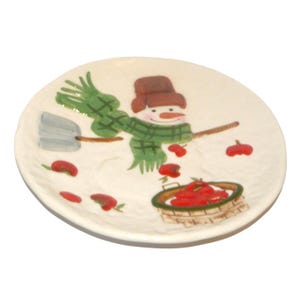 May include: A round, off-white ceramic plate featuring a whimsical snowman design. The snowman wears a brown hat and a green scarf, holding a broom. Red apples are scattered around a basket filled with more apples, creating a festive, seasonal theme.
