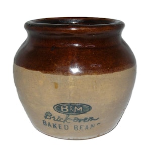 May include: A round ceramic bean pot with a two-tone glaze featuring a dark brown upper section and a light tan lower section. The front displays the text B&M inside an oval, followed by the words Brick-oven BAKED BEANS in dark blue lettering.