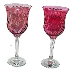 Puede incluir: Dos copas de vino color arándano con un cuenco en forma de tulipán y un tallo transparente y retorcido. Las copas tienen un borde ondulado y se asientan sobre una base circular transparente. El cristal tiene un diseño en espiral.