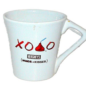 May include: White ceramic mug with a unique handle design. The mug features the Hershey's logo and the words "xoxo" in red, with a red Hershey's Kiss graphic. The text "(HUGS & KISSES.)" is also printed on the mug.