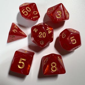 May include: Seven red dice with gold numbering are scattered on a white surface. The dice are various shapes, including a d20, d10, d8, d6, and a d4. The numbers are clearly visible, ranging from 1 to 50.