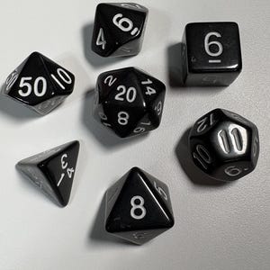 Puede incluir: Un conjunto de siete dados poliédricos negros con números blancos. Los dados incluyen una variedad de formas, como un d4, d6, d8, d10, d12, d20 y un d50. Perfectos para juegos de rol y de mesa.