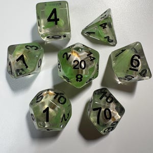 May include: Set of seven clear, multi-sided dice with green and white inclusions. Each die has black numbers. The dice are designed for tabletop role-playing games.