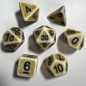 May include: A collection of seven ivory-colored dice with black numbering. The dice are various shapes, including a four-sided die, a six-sided die, an eight-sided die, a ten-sided die, a twelve-sided die, a twenty-sided die, and a percentile die. The dice are for tabletop games.
