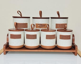 May include: A set of seven cylindrical ceramic canisters with speckled white bodies and brown bases, arranged on a wooden rack. Each lid features a brown loop handle. The canisters are labeled with embossed text reading Sugar, Tea, Coffee, and Spice.