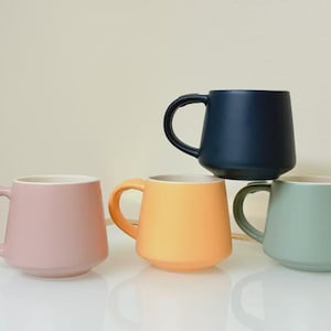 May include: Four ceramic mugs in pastel colors: pink, orange, green, and navy blue. Each mug has a rounded shape with a handle and a white interior. The mugs are stacked and arranged on a white surface.