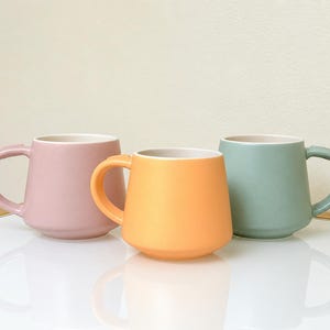May include: Three ceramic mugs with a matte finish, each featuring a rounded body and a curved handle. The mugs are colored in soft pastel shades of dusty pink, vibrant orange, and muted sage green, all with a white interior and base.
