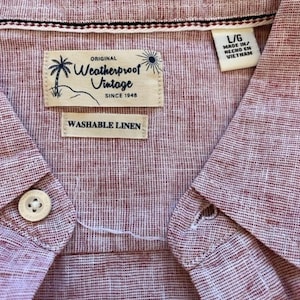May include: Close-up of a red and white striped linen shirt. The shirt has a button-down collar and a button closure. The label reads "Weatherproof Vintage" and "Washable Linen". The shirt is a size L/G and made in Vietnam.