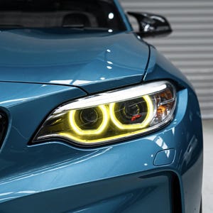 May include: A teal sports car with bright yellow headlights. The front of the car is visible, showcasing the grill and the sleek design of the vehicle. The headlights are illuminated, adding a striking visual element.