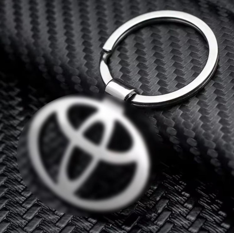 May include: A silver-colored Toyota logo keychain. The keychain features a circular key ring and a round charm with the Toyota emblem. The background is a dark, textured surface.