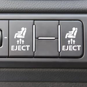 May include: A close-up of a grey car dashboard featuring white symbols. The image shows two buttons with a seat icon, arrows, and the word "EJECT" in white. The buttons are located on the centre console.