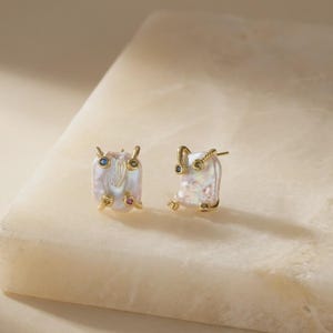 May include: A pair of square-shaped pearl stud earrings with gold accents. Each earring features a large, irregular white pearl with gold-colored metal detailing and small, colorful gemstones. The earrings are displayed on a light-colored surface.