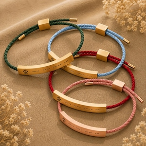May include: Four adjustable bracelets featuring gold-toned curved metal bars. Each bar is engraved with a paw print and Morse code-style dots and dashes. The bracelets have braided cords in dark green, light blue, red, and pink, with gold-toned sliders.