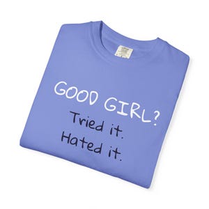 Puede incluir: Una camiseta azul bígaro doblada con el texto "GOOD GIRL? Tried it. Hated it." en blanco y negro. La camiseta tiene una etiqueta en la esquina superior derecha.