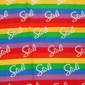 以下が含まれることがあります： 白の筆記体で「Stoli」の文字が繰り返された、虹色のストライプの織物。赤、オレンジ、黄色、緑、青、紫の水平バンドが特徴です。織物は長方形です。