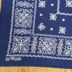May include: A navy blue bandana with a white paisley design. The bandana features a repeating pattern of floral and scrollwork motifs. The lower left corner has the text "FAST COLOR 100% COTTON".