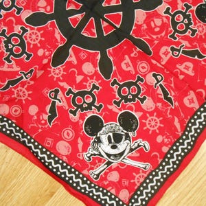 Puede incluir: Bandana roja con temática pirata con borde en blanco y negro. La bandana presenta un timón negro, calaveras, espadas cruzadas y un personaje de ratón de dibujos animados con parche en el ojo y sombrero de pirata.