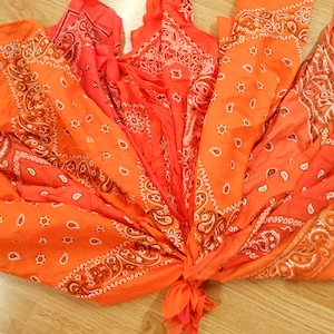 Puede incluir: Arreglo de pañuelos naranjas y rojos, desplegados para mostrar sus diseños de cachemira. Los pañuelos están atados en la base, creando una presentación decorativa y vibrante. Los pañuelos tienen detalles blancos y marrones.