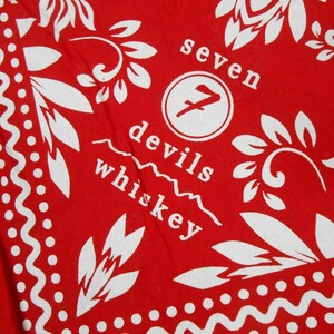 Può includere: Bandana rossa con disegni floreali e a pois bianchi. Il centro presenta il testo "seven devils whiskey" in un font stilizzato, con un logo di un "7" stilizzato all'interno di un cerchio. La bandana ha un bordo decorativo.
