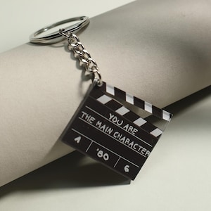 May include: A black and white film clapperboard keychain with the text "YOU ARE THE MAIN CHARACTER". The keychain has a silver chain and ring. The clapperboard has the numbers "A '80 6".