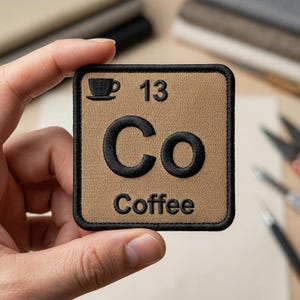 May include: A square, tan patch with black border, featuring a coffee cup icon, the number 13, and the word "Coffee" below the letters "Co". The patch is held in a hand.