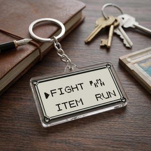 May include: Clear rectangular keychain with the words "FIGHT", "ITEM", and "RUN" in a pixelated font. The keychain is attached to a silver keyring and chain. The background includes keys, a pen, and a notebook.