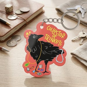 May include: A red keychain with a cartoon raven illustration and the text "Collector of Trinkets." The raven is black with a gold necklace and various trinkets around its feet. A silver key ring and chain are attached.