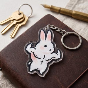 May include: A clear acrylic keychain featuring a cartoon rabbit flexing its muscles. The rabbit is white with pink accents and a black outline. The keychain is attached to a silver chain and ring. Keys and a pen are in the background.